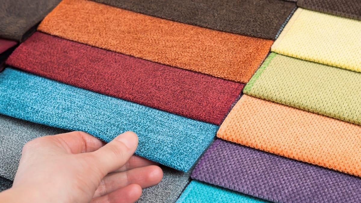 A hand lifting a blue textile sample from a stack of colorful swatches to inspect the fabric grain and feel