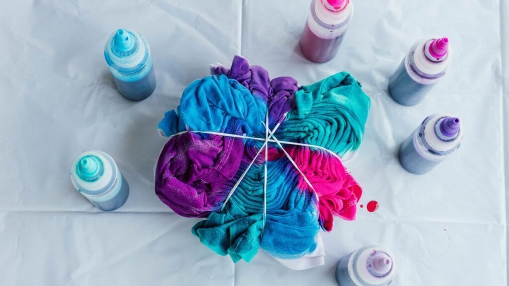 A tie dye shirt bundled and tied with rubber bands with dye bottles around it, featuring bright purple, teal, and magenta colors