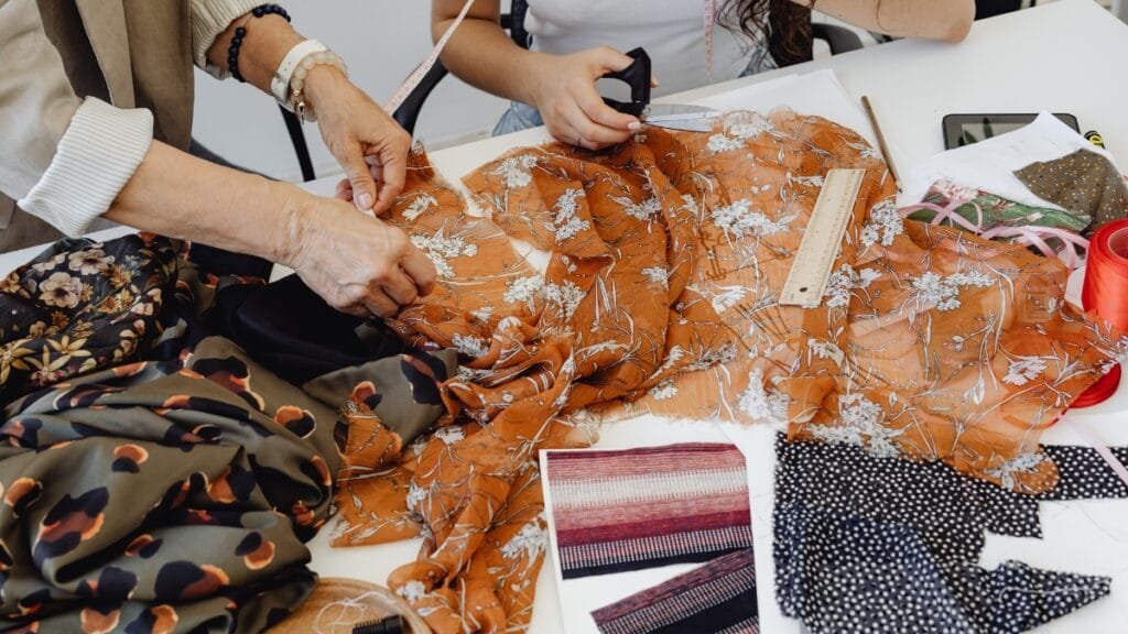 A person cuts fabric with scissors on a table, surrounded by fabric swatches, showing the process of working with different materials for a design