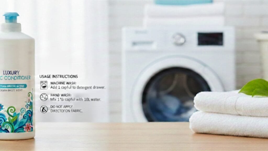 A bottle of fabric conditioner sits on a wooden surface next to a neatly folded stack of white towels. In the background, a washing machine is visible. A sheet with usage instructions is placed beside the bottle. The label on the bottle includes the word “LUXURY” and “C CONDITIONER,” with blue and teal decorative swirls on a white bottle with a blue cap.