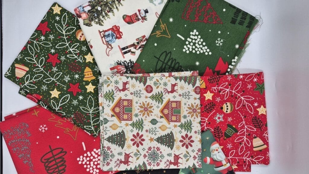 A curated selection of Christmas fabrics laid out by the yard, including velvet, flannel, cotton, and satin in red, green, gold, and ivory with patterns like plaid, snowflakes, and holly.