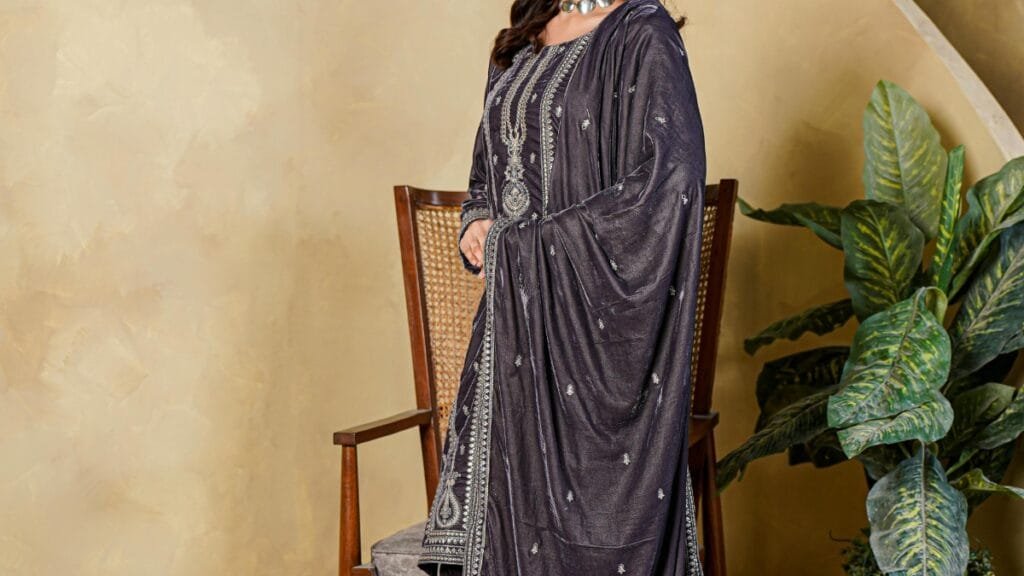 A woman standing indoors wearing a dark traditional embroidered outfit with a jeweled neckline, posed beside a chair and potted plants.
