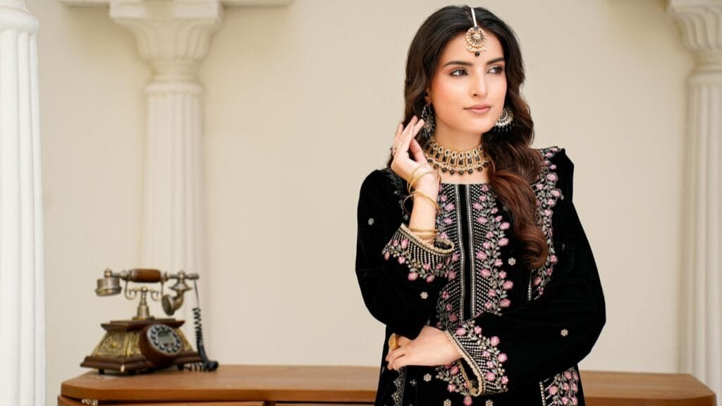 Elegant woman in a black embroidered traditional outfit with pink floral detailing, layered gold jewelry, and a maang tikka, posing indoors beside a vintage telephone.