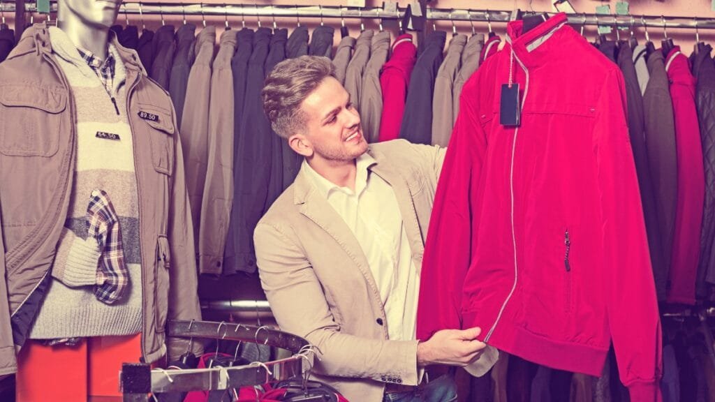 A man in a clothing store inspects a bright pink jacket on a rack, surrounded by other hanging coats and a striped price tag.