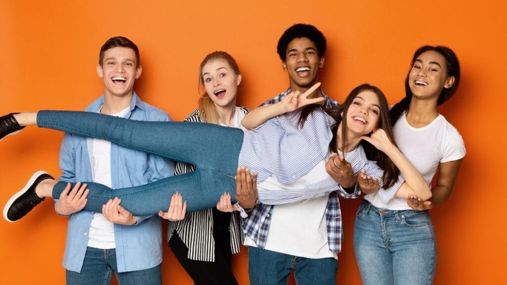 A group of five young people pose and laugh together in front of an orange background, wearing denim jeans and casual tops.