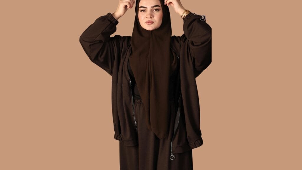  A woman in a brown abaya and beige hijab, holding a beige handbag, with sunglasses on, standing against a neutral background.