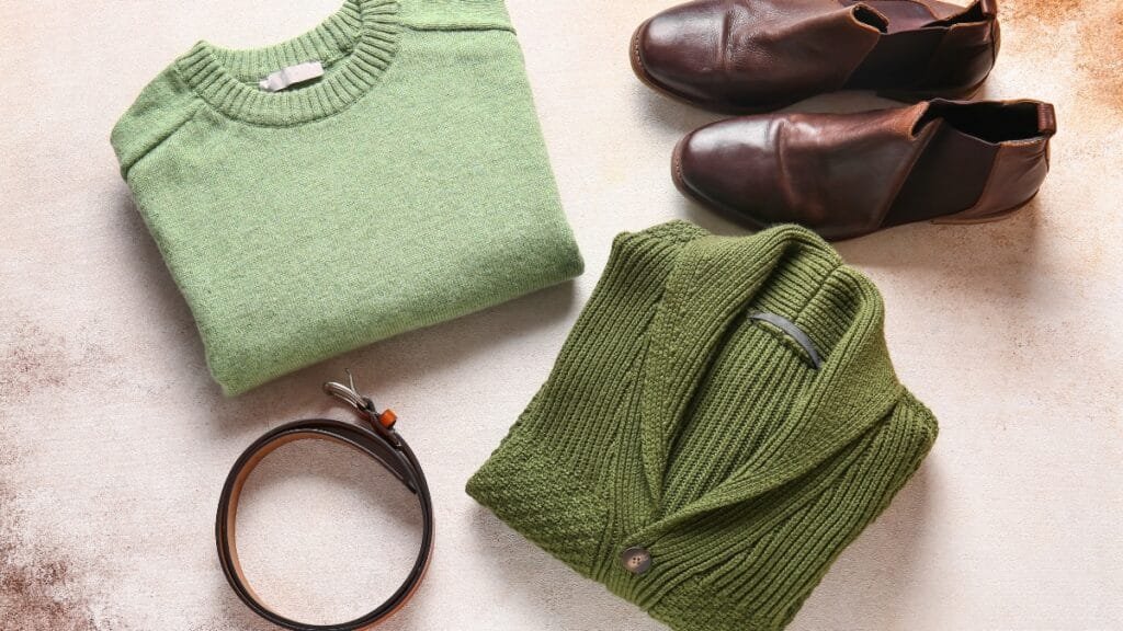 A flat lay of a folded green knit sweater, another green ribbed sweater with brown buttons, a brown leather belt, and a pair of brown leather shoes on a light beige surface.