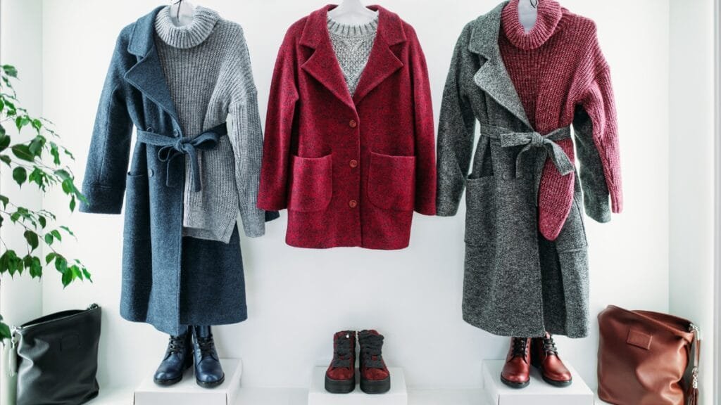 A rack of three folded sweater-and-skirt outfits on display: blue, burgundy, and gray wool garments with matching belts, paired with boots, with shoes arranged on a white shelf against a plain light wall