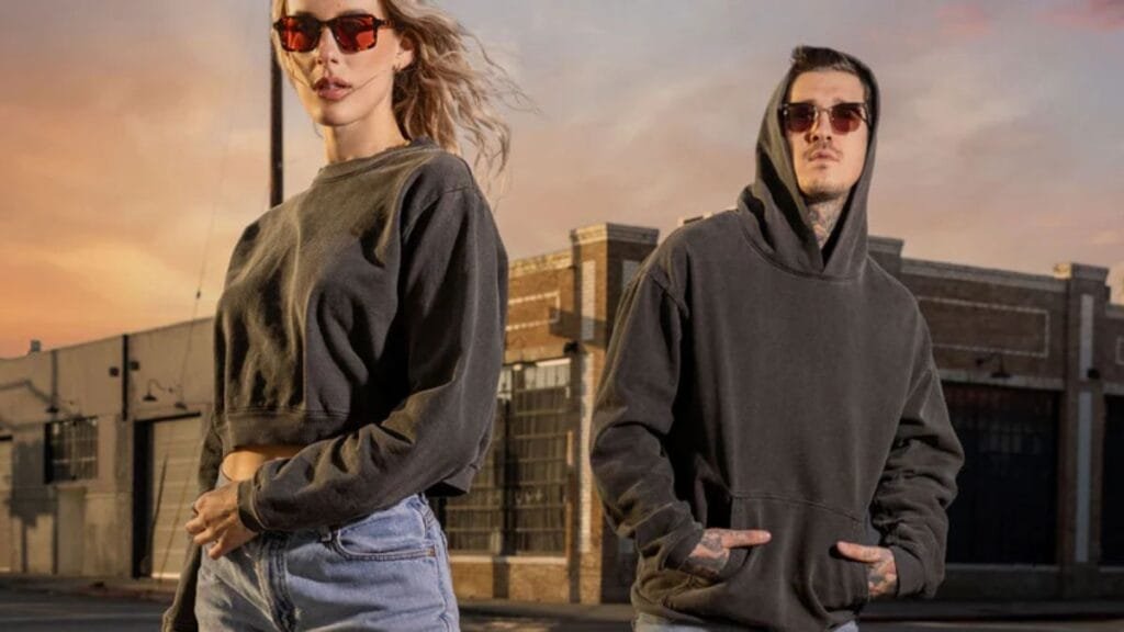 Two models wearing casual streetwear, including hoodies and sunglasses, standing outdoors in an urban setting at sunset.