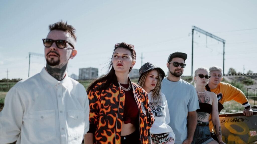 Group of young adults in casual streetwear, including jackets, hats, and sunglasses, standing outdoors near railway tracks and looking off into the distance.