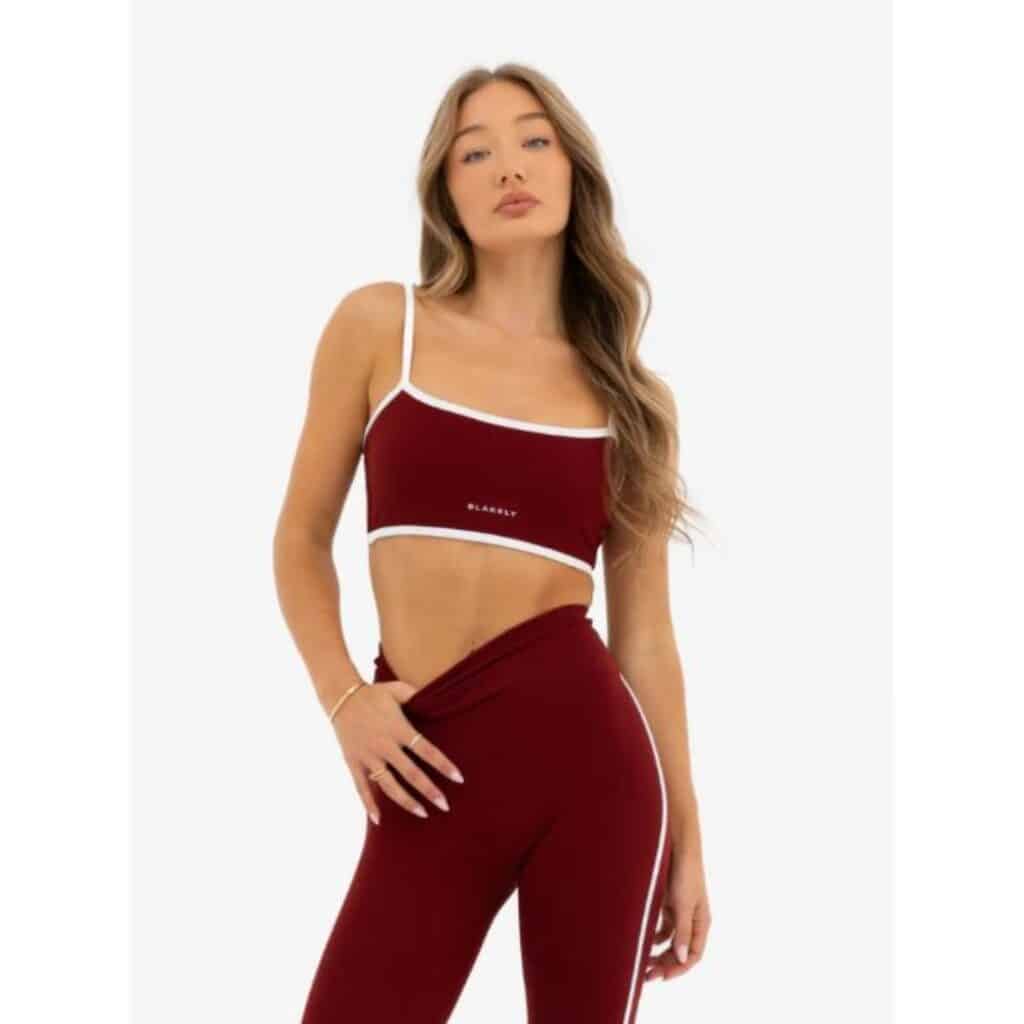 A woman poses in a wine-colored matching crop top and leggings workout set with white trim and side stripes, standing against a plain white background.