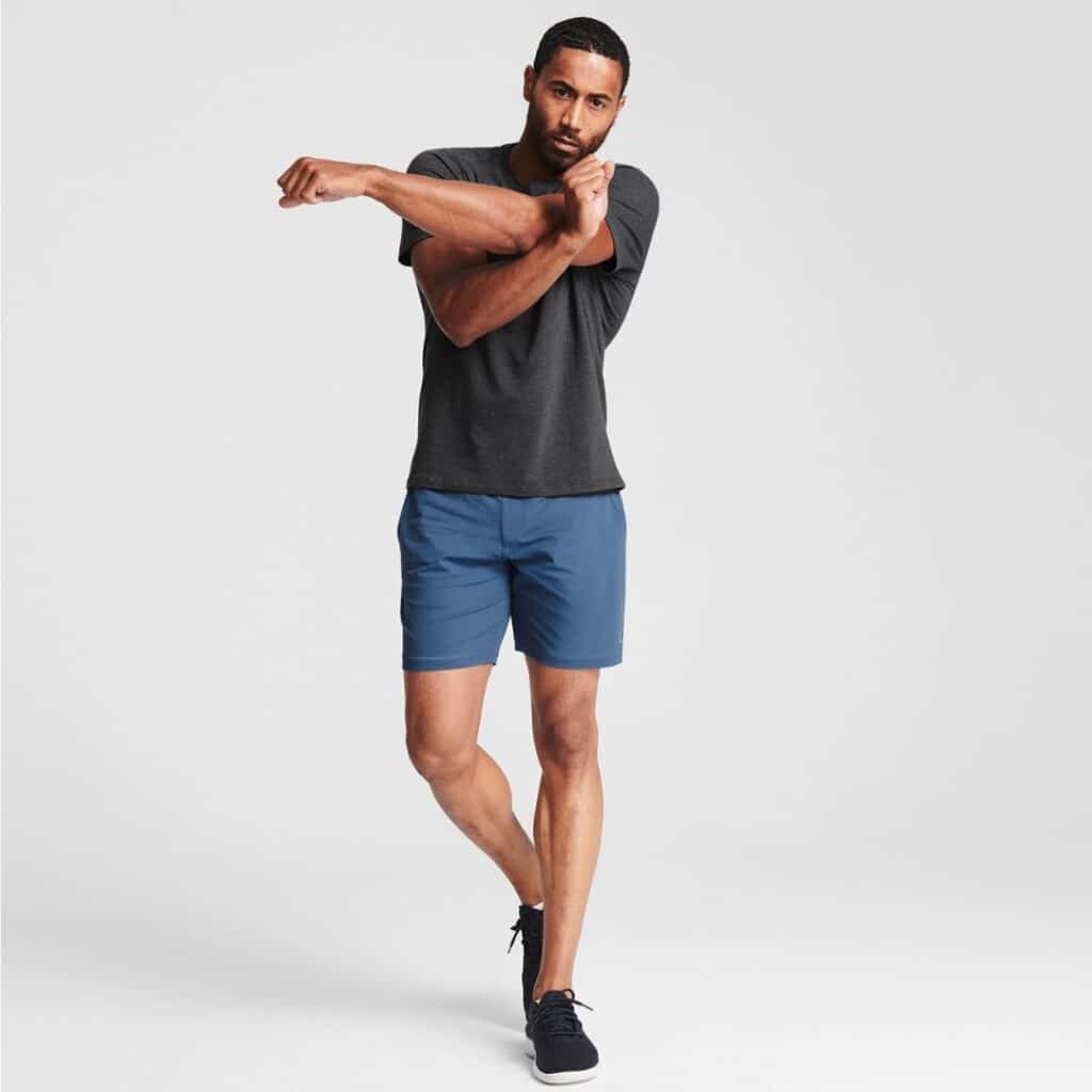 Man wearing a gray athletic t-shirt and blue shorts stretching his arm on a white background.