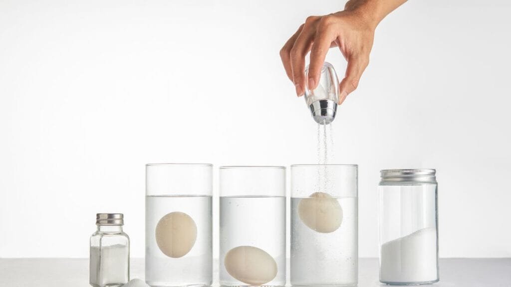 Hand sprinkling salt into glasses of water with eggs, demonstrating density and liquid behavior in a simple experiment