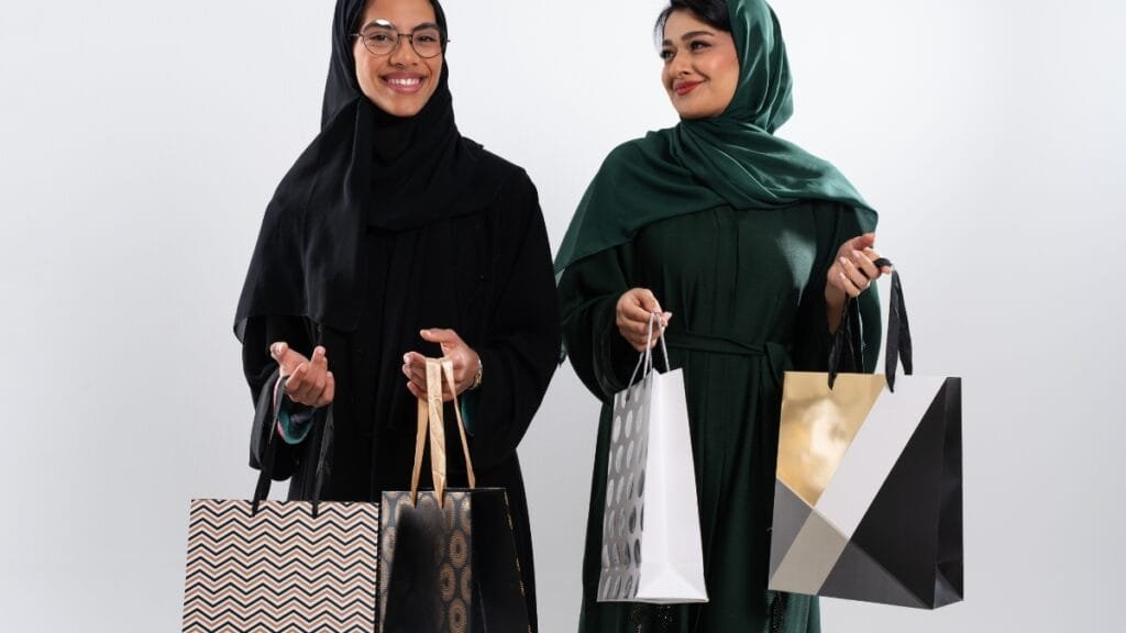 Two women in abayas and hijabs, smiling and holding shopping bags.