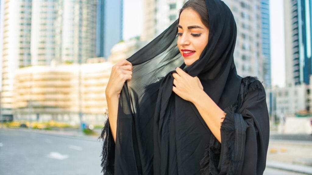 Woman wearing a black abaya and headscarf, smiling and adjusting her scarf in an urban setting.