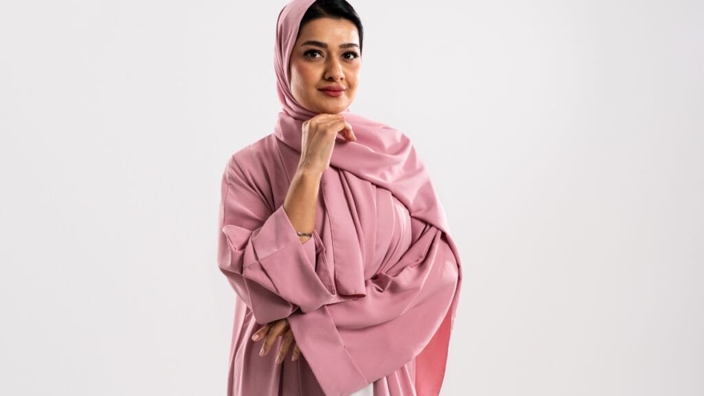 A woman in a pink abaya and hijab, posing thoughtfully with her hand on her chin.