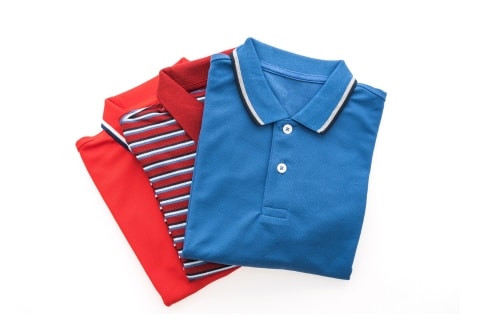 Three folded polo shirts stacked on a white surface — blue with striped collar on top, striped red in the middle, solid red on the bottom.