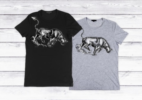 Two graphic T-shirts, one black and one gray, laid side-by-side on a white wooden surface, each featuring a bull graphic.
