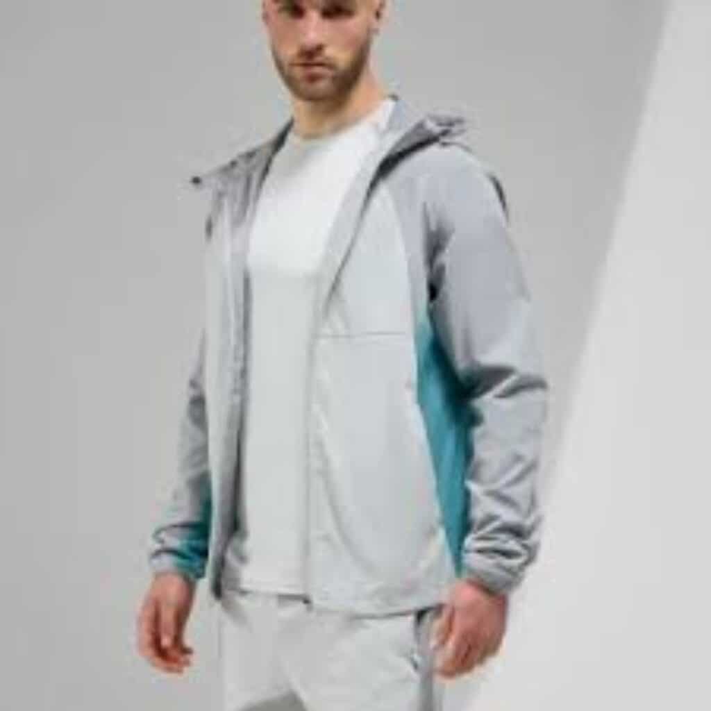 A man in a grey-and-light-blue hooded athletic jacket and white pants standing against a white background.
