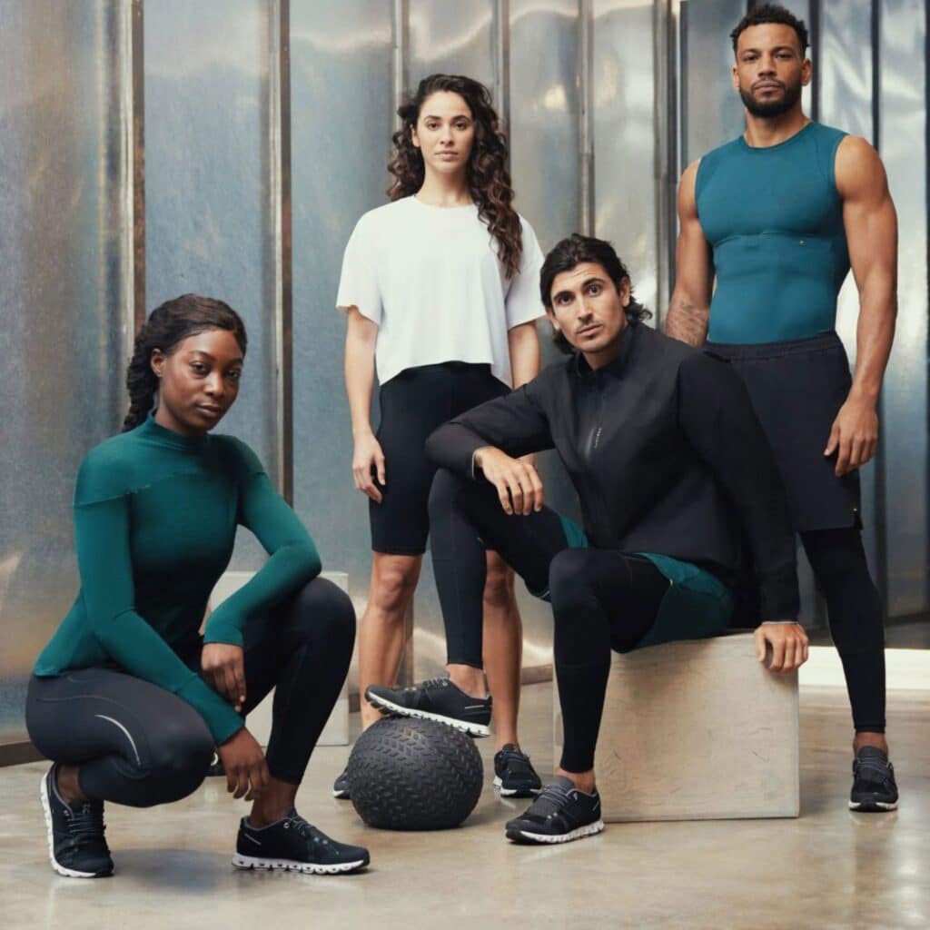 Group of four athletes — two women and two men — posing in a gym with a textured metal wall background and a medicine ball in the foreground.