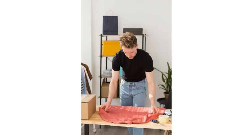 A man standing at a wooden table folding a coral-colored garment, with a cardboard box to his left and crafting supplies like scissors and tape to his right. A shelving unit and potted plant are visible in the background.