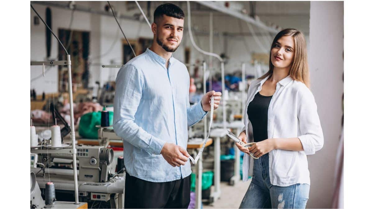 A man and woman standing in a garment factory, holding a measuring tape and scissors, with sewing machines and spools of thread in the background.