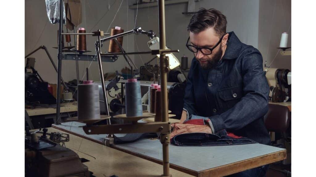 A bearded tailor in a denim shirt sewing dark fabric with red lining at a workshop sewing machine.