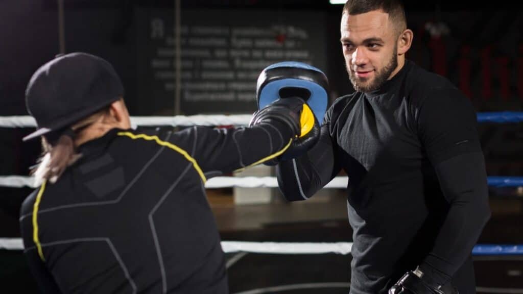 A boxing trainer holds a blue and yellow punch mitt for a boxer in a dark gym with a boxing ring in the background. The trainer, wearing a black long-sleeved shirt, looks directly at the boxer who is out of frame. The boxer's arm, clad in a black and yellow long-sleeved shirt and a black boxing glove, is extended, making contact with the mitt. The boxing ring ropes are visible, appearing white or light blue against the dark background.