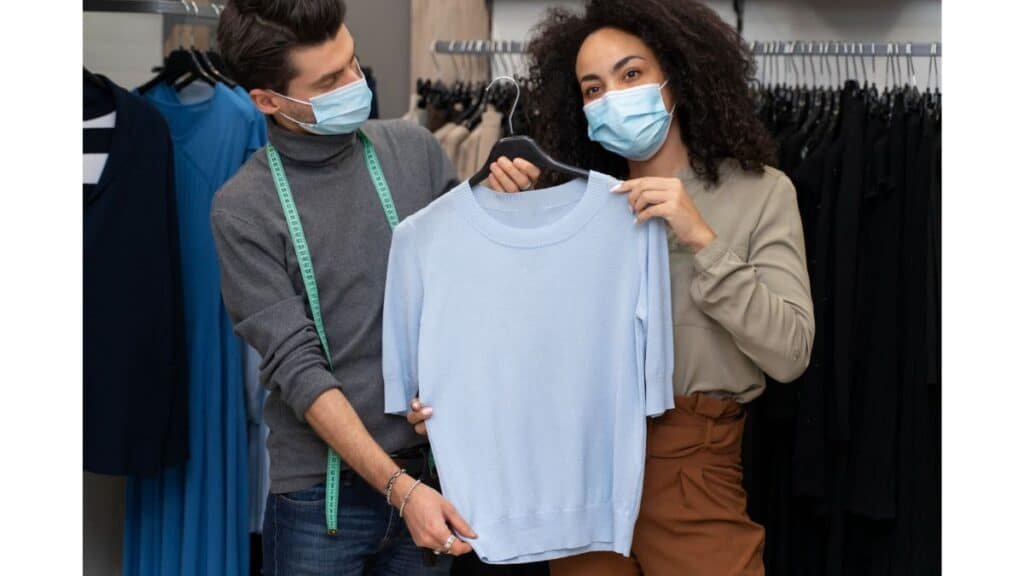 Two people in surgical masks holding up a light blue sweater on a hanger in a clothing boutique.