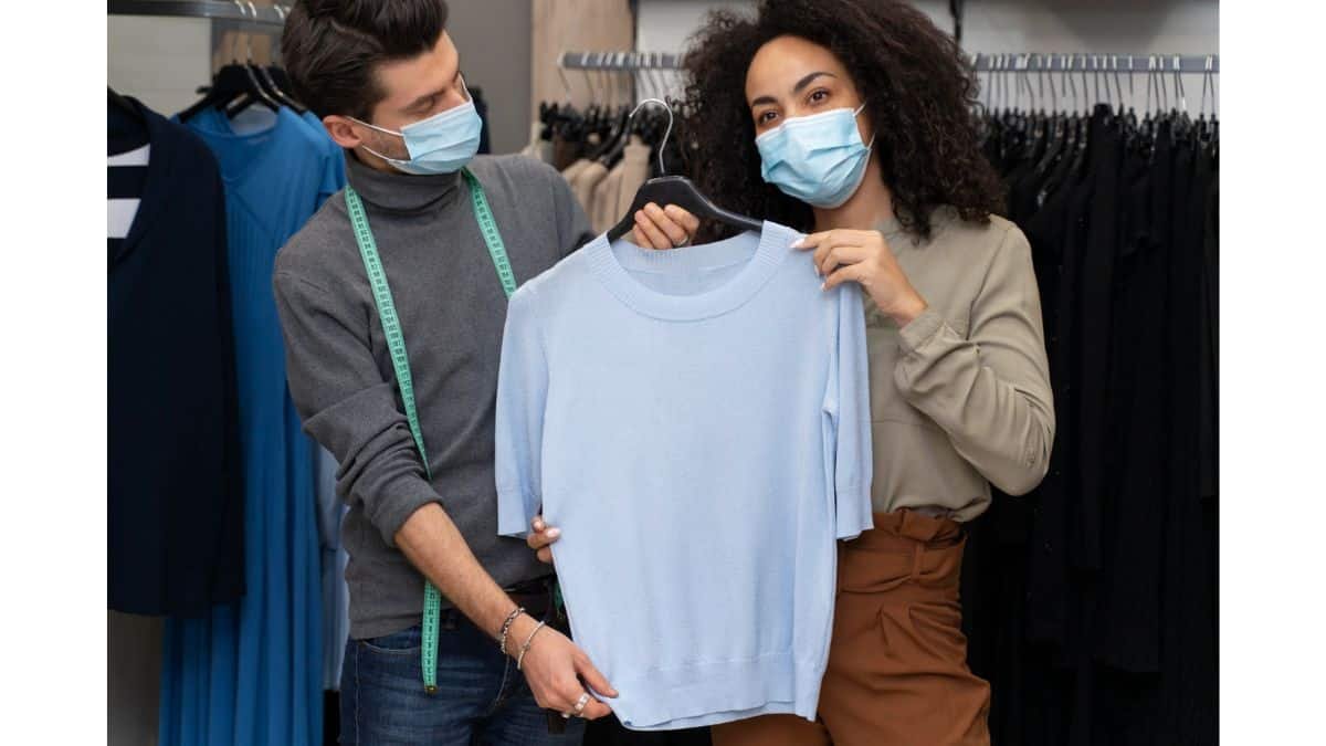 Two people in surgical masks holding up a light blue sweater on a hanger in a clothing boutique.