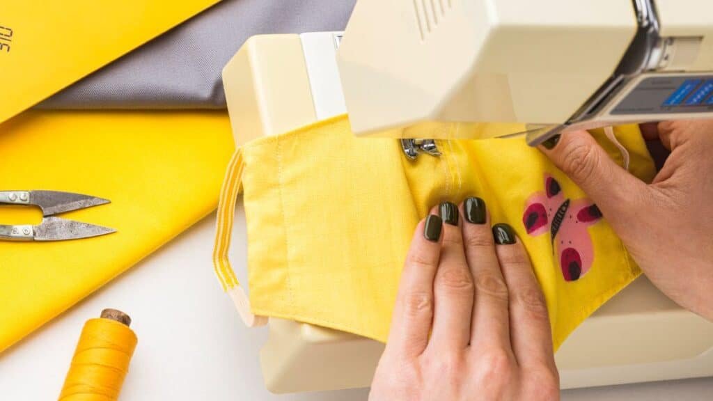 A close-up overhead shot of hands sewing a yellow fabric face mask with a red and pink butterfly pattern on a beige sewing machine. The person's fingernails are painted dark green. To the left of the machine, fabric scissors rest on a bright yellow fabric piece partially covered by gray and yellow fabric, with a spool of orange-yellow thread below. The background is mostly white.