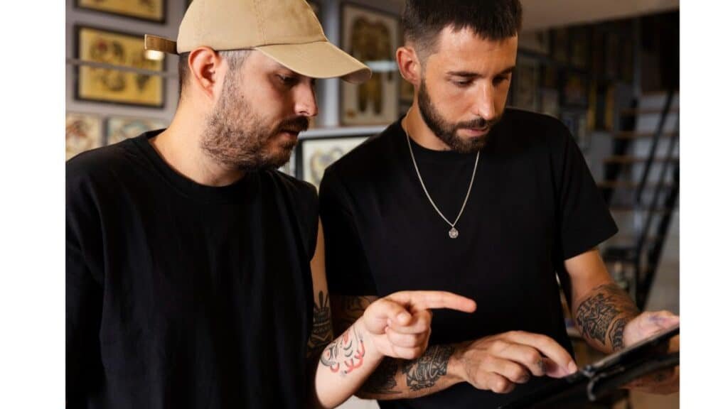 Two men with beards and short hair stand side-by-side wearing black t-shirts. The man on the left wears a beige baseball cap backward. The man on the right wears a silver chain with a round pendant and points off-camera toward the lower right. Both have tattooed forearms. The background is slightly blurred with framed images visible on a wall.