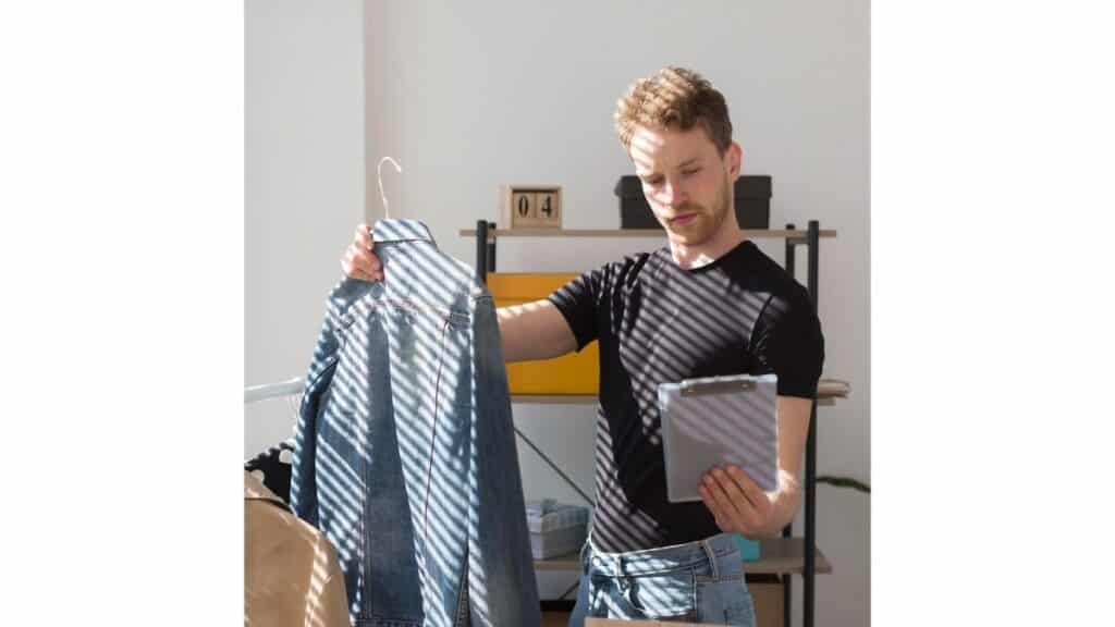 A man with short brown hair and a beard, wearing a black t-shirt and jeans, holds a denim jacket on a hanger while looking down at a tablet. Diagonal shadows fall across the scene from a window.