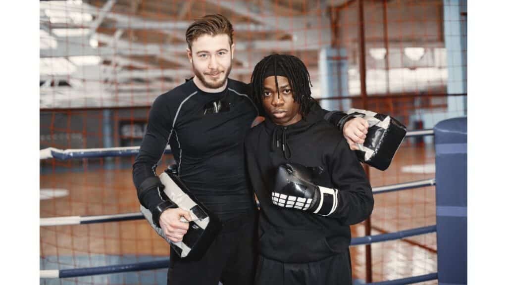 Two coaches stand side-by-side in a boxing ring, each holding a boxing mitt.