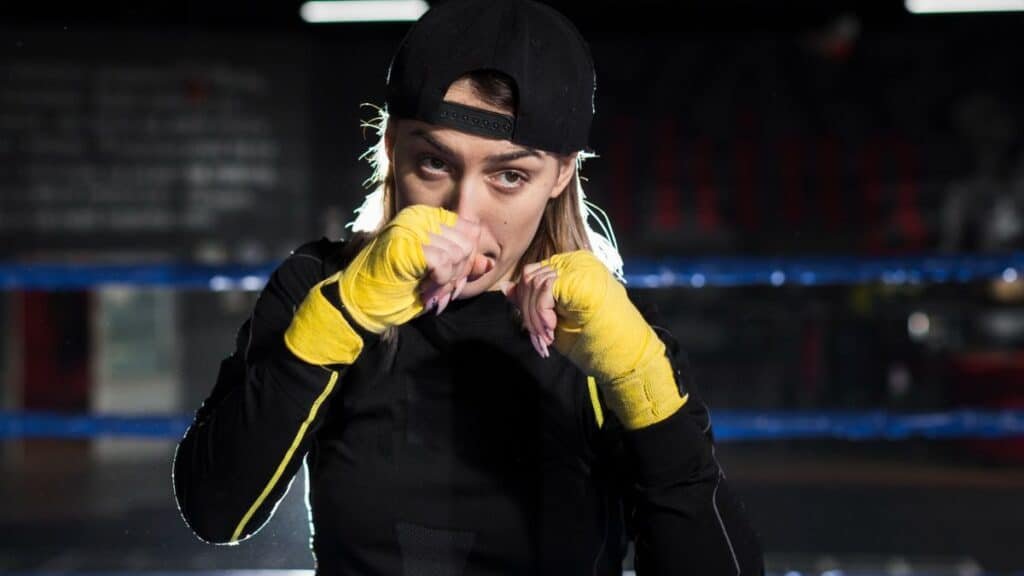 A focused female boxer stands in a dimly lit boxing ring, her face partially obscured by her wrapped hands. She wears a black long-sleeved shirt and a backward black baseball cap, with her hands wrapped in bright yellow boxing tape and light pink fingernails visible beneath the wraps. Her light brown hair is highlighted by backlighting. The blurred background shows hints of blue ropes and red arena seating.