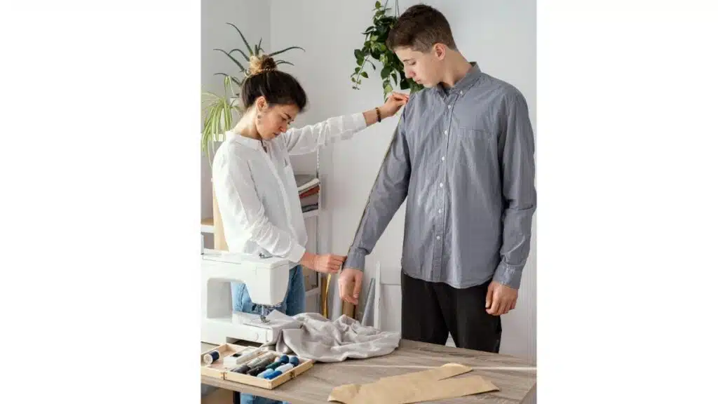 A woman measures a man's sleeve with a yellow tape measure in a sewing workspace with a sewing machine, fabric swatches, and thread.