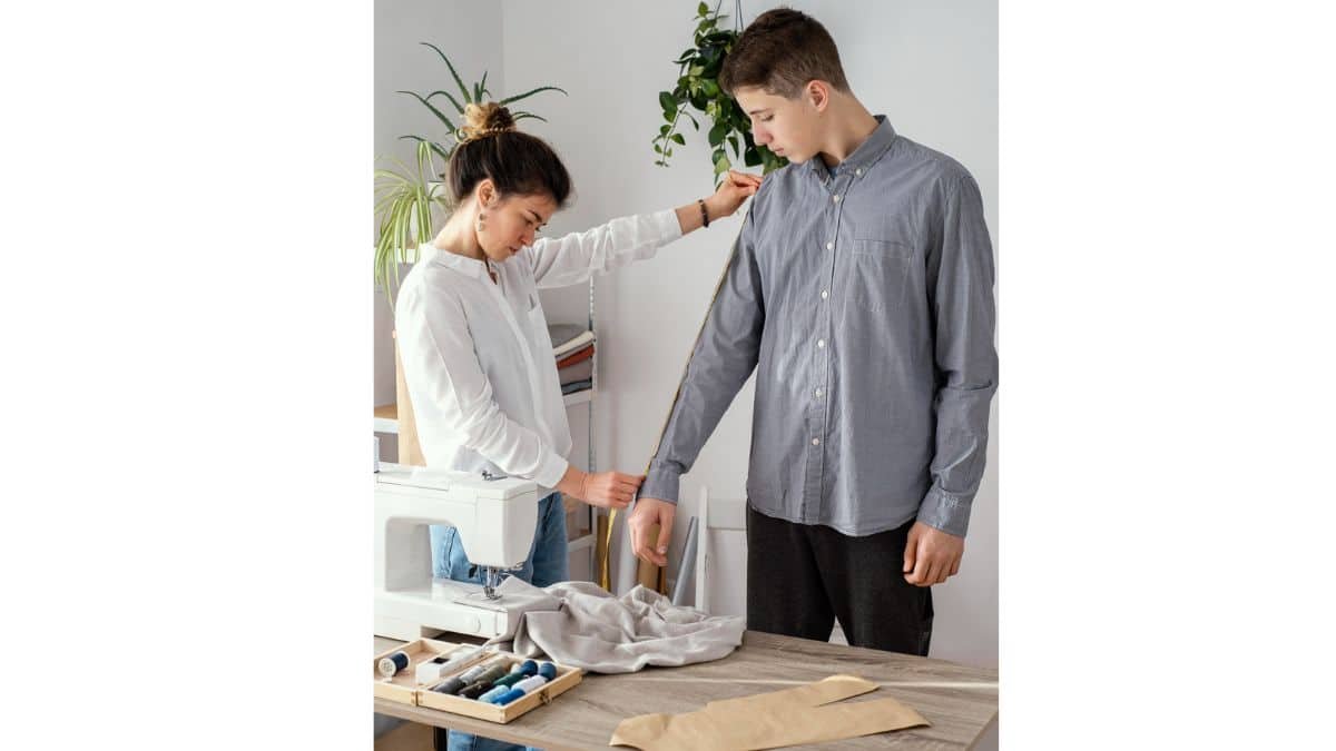 A woman measures a man's sleeve with a yellow tape measure in a sewing workspace with a sewing machine, fabric swatches, and thread.