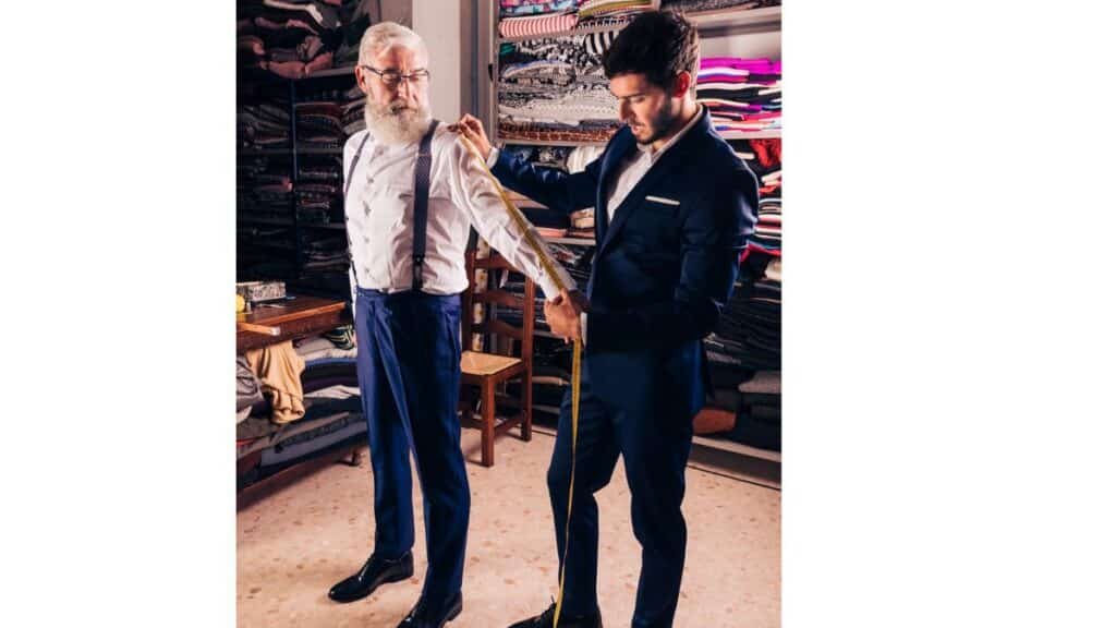 A tailor in a suit measures a customer's arm in a fabric shop.