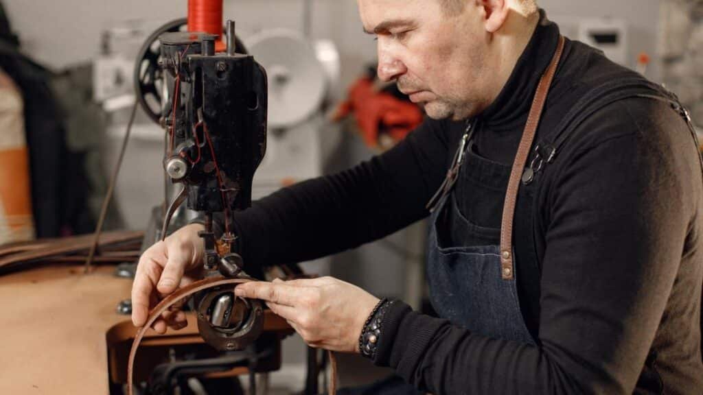 A man in a denim apron sewing brown leather on a vintage industrial machine with red thread.