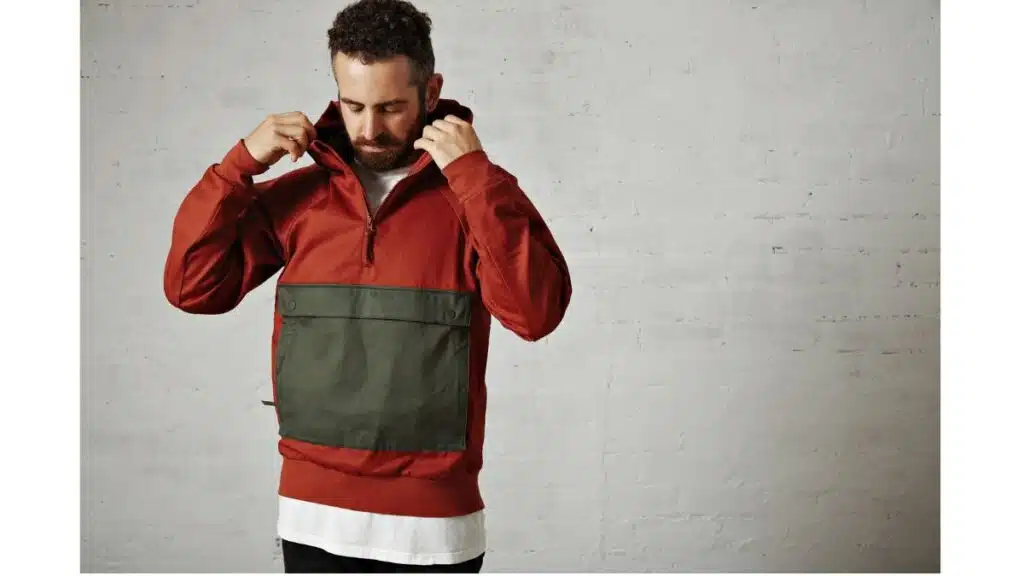 A bearded man with curly dark hair adjusts the hood of his rust-colored anorak while standing against a white brick wall.