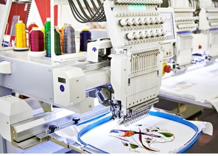 A sewing machine at a craft or textile workstation with threads, fabrics, and supplies in the background.