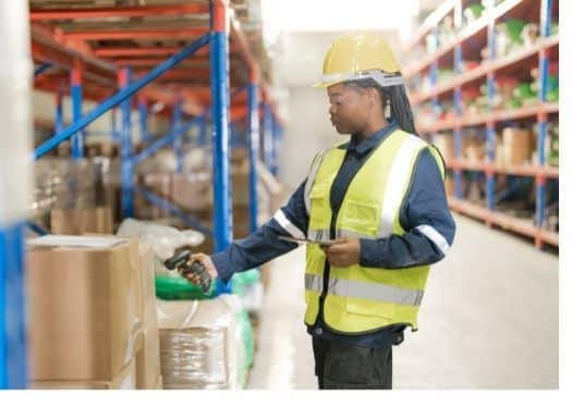 Worker in a warehouse wearing a hard hat and reflective vest, inspecting a product aisle with a handheld device.