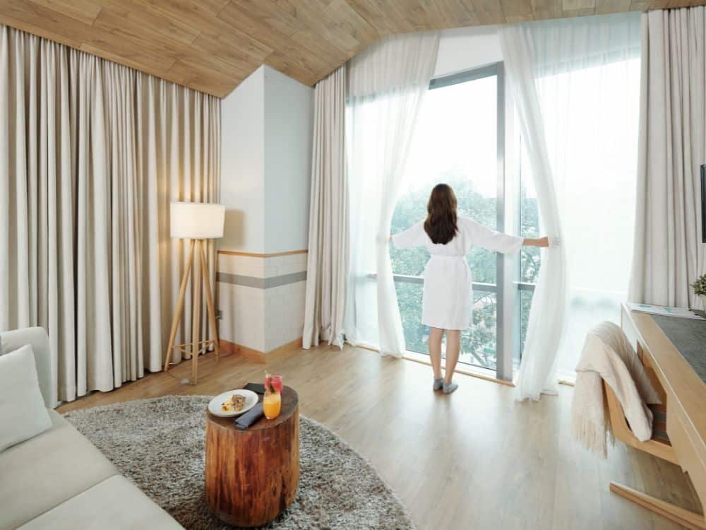 A woman in a bathrobe opening sheer curtains in a modern hotel room with a cozy atmosphere.