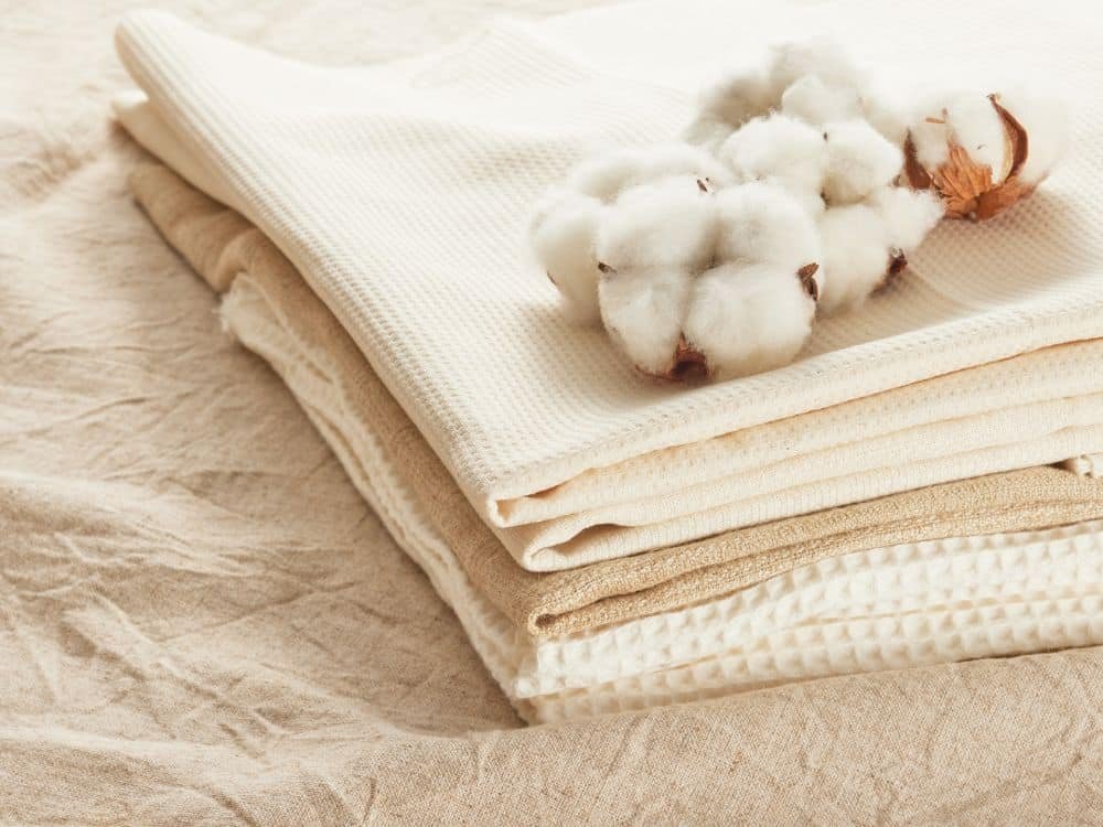 A neatly arranged stack of neutral-colored fabrics with a sprig of cotton on top, showcasing the softness and texture of the materials.