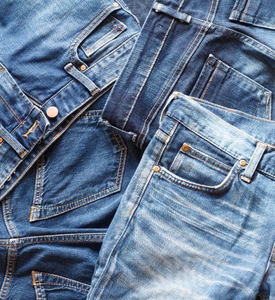 Overhead close-up of a pile of blue jeans in various shades of indigo and light blue, showing different denim washes, textures, and styles.