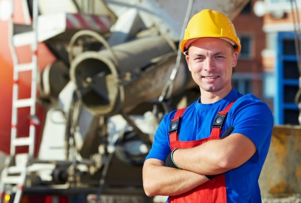 A construction worker wearing a yellow safety helmet and red overalls, standing with arms crossed and smiling at the camera on a construction site.