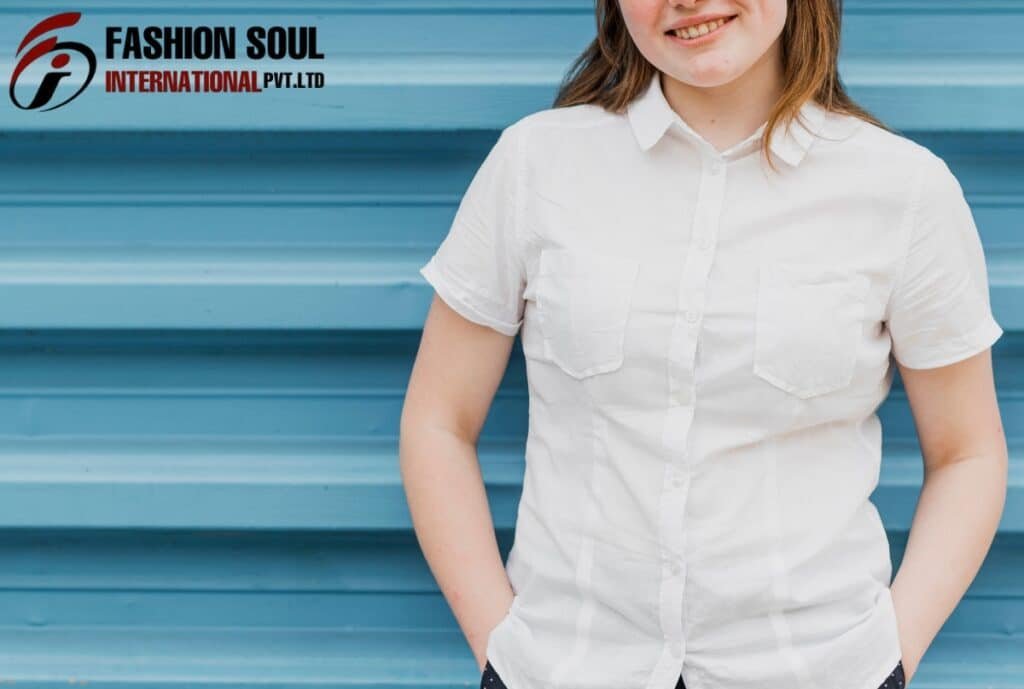 A woman with light brown hair, wearing a white short-sleeve button-up shirt, stands in front of a blue, horizontally-striped background with her hands in her pockets, smiling slightly.