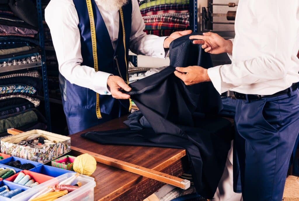Two men are handling a piece of dark fabric in a tailoring or fabric shop, with shelves of folded cloth and sewing supplies around them.