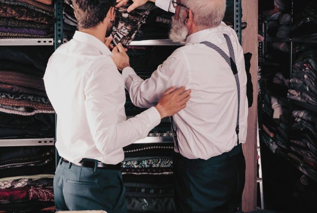 Two men engaged in a discussion, one younger and one older, in a fabric or clothing store. They are both wearing white shirts and dark trousers, with the older man also wearing suspenders. They are standing in front of shelves filled with folded textiles or clothing items, pointing and examining fabric.