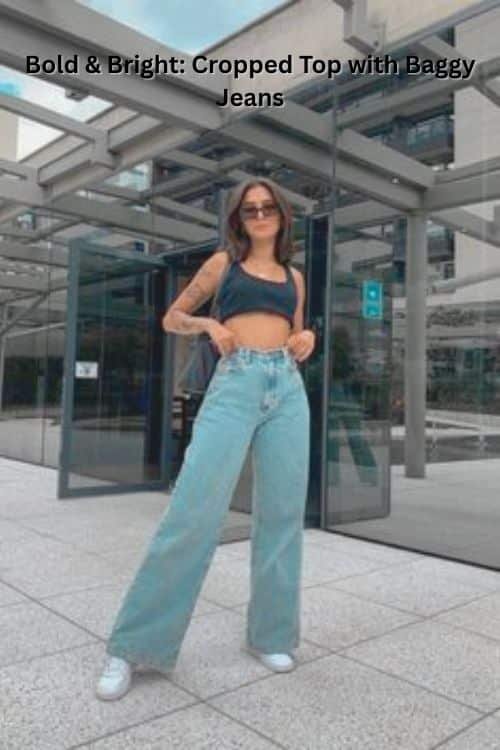 A woman stands outdoors in front of a modern glass building, wearing a black cropped top, high-waisted light blue wide-leg jeans, white sneakers, and sunglasses.
